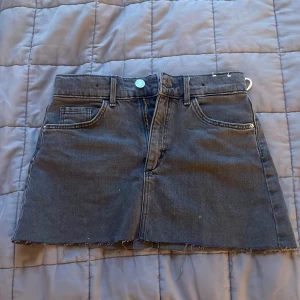 Svart jeanskjol från H&M - Svart jeanskjol från H&M med klassisk femficksdesign och rå kant nertill. Kjolen har dragkedja och knapp framtill samt bälteshällor. Perfekt för en cool och avslappnad stil.