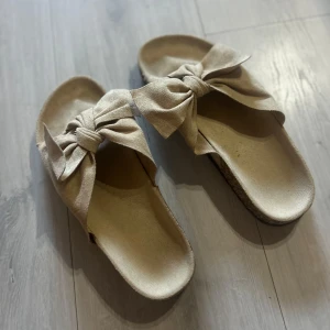 Beige sandaler med rosett - Säljer ett par snygga beige sandaler med stora rosetter framtill. Perfekta för sommaren och lätta att bara glida i. De har platt sula och öppen tå, vilket gör dem både bekväma och luftiga.