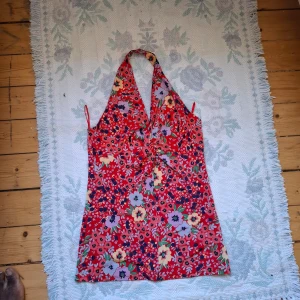 Röd blommig halterneckklänning - Säljer en färgglad röd halterneckklänning med blommigt mönster i lila, gult och grönt. Klänningen har omlottkänsla framtill och knyts i nacken. Perfekt för varma dagar och somriga tillfällen.