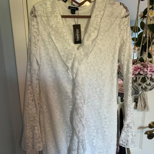 Vit spetsklänning med volanger och lång ärm från Boohoo - Säljer en vit spetsklänning från Boohoo med v-ringning och långa ärmar. Klänningen har ett romantiskt blommönster i spets och är i minilängd. Perfekt för dig som vill ha en feminin och elegant look. Klänningen är något liten i storlek skulle säga 42/44. Den är lite för kort för mig som är 179