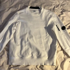 Vit tröja från C.P. Company - Säljer en stilren vit sweatshirt från C.P. Company med rund hals och ribbade muddar. Tröjan har en ikonisk svart linsdetalj på ena ärmen och är tillverkad i mjuk bomull. Perfekt för en clean och modern look. Använd 1 gång få nu den är för liten. Passar också M, köpt för 1800kr