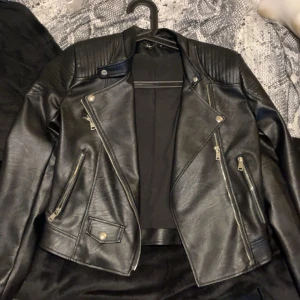 Svart skinnjacka från SHEIN - Säljer en svart skinnjacka från SHEIN i storlek XS. Jackan har klassisk bikerstil med sned dragkedja, flera fickor med dragkedjor och detaljerade axelpartier. Perfekt för dig som vill ha en edgy look.