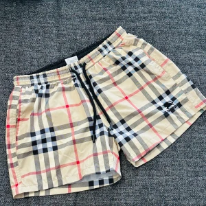 Rutig shorts från Burberry - Snygga rutiga shorts från Burberry i klassiskt beige, svart och rött mönster. Shortsen har elastisk midja med snörning och två fickor framtill. Perfekta för sommaren och har en broderad Burberry-logga på ena benet.