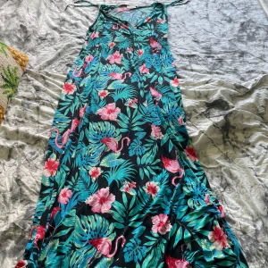 Blommig långklänning med flamingos - Säljer en färgglad långklänning med tropiskt blommönster och flamingos. Mönstret går i grönt, rosa och blått mot en mörk bakgrund. Superfin till stranden eller picknick! Ha ringen koll på storleken men runt 40-50