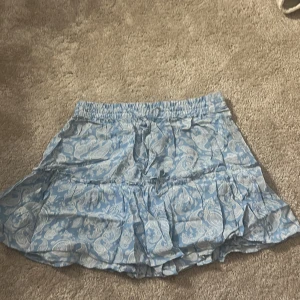 Blåvit mönstrad kortkjol från Zara - Supersöt blå och vit kortkjol från Zara med paisleymönster och volangdetaljer. Kjolen har resår i midjan och är luftig och lätt, perfekt för varma dagar. Säljer då den inte kommer till användning längre! Är inbyggda shorts