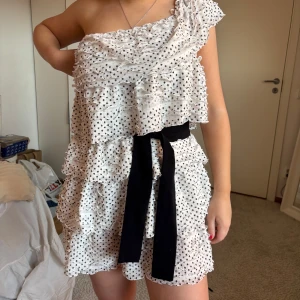 Vit prickig omlottklänning från Zara - Supersöt vit kortklänning från Zara med svarta prickar, volanger och en axel. Klänningen har omlottkänsla och ett brett svart knytband i midjan som ger extra stilpoäng.