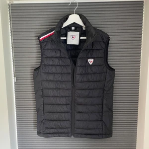 Svart dunväst från Rossignol - svart dunväst från Rossignol med quiltad design och dragkedja framtill. Västen har märkets logga på bröstet och en röd, vit och blå detalj på axeln. Storlek L. Mycket fint skick.