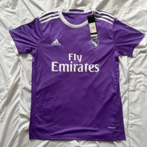 Lila Real Madrid fotbollströja Ronaldo 7 - Säljer en lila Real Madrid fotbollströja från Adidas med Ronaldo 7 på ryggen. Tröjan har vita detaljer, rund hals och klassiska Adidas-ränder på axlarna. Fly Emirates tryck på bröstet och klubbmärke på vänster sida.