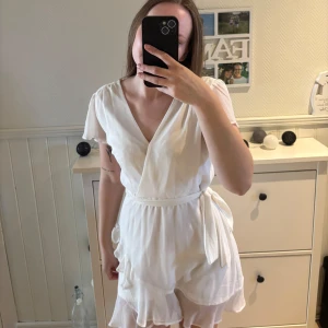 Vit omlottklänning med volang - Säljer en söt vit omlottklänning med korta ärmar och volangdetaljer längst ner. Klänningen har knytband i midjan och en lätt v-ringning. Storlek M men sitter mer som en S. Perfekt för student och sommaren