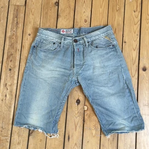 Randiga jeansshorts från Replay - Säljer ett par blå och vita randiga jeansshorts från Replay. Klassisk femficksmodell med knappgylf och råa benslut. Perfekta för sommaren och har en avslappnad passform.