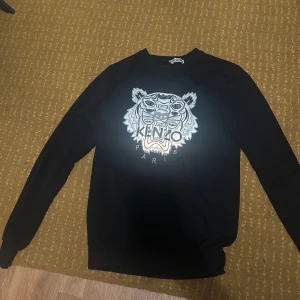 Svart sweatshirt med tigertryck från Kenzo - Säljer en svart sweatshirt från Kenzo med det ikoniska tigerhuvudet och texten 'Kenzo Paris' på bröstet. Tröjan har rund hals och långa ärmar, perfekt för en cool och avslappnad stil. Reflekterande märke i mörkret!