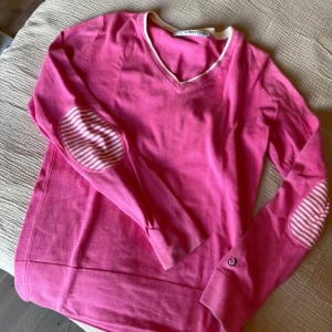 Rosa  tröja från Lobster Golfwear - Säljer en rosa stickad tröja från Lobster Golfwear med vita detaljer och randiga armbågslappar. Tröjan har v-ringning och långa ärmar. 