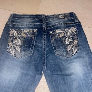 Blå jeans med broderade bakfickor från Miss Me - Säljer ett par blå jeans från Miss Me med snygga broderade detaljer och stenar på bakfickorna. Jeansen har bootcut-modell. Och är i fint skick, jag sydde in ett resårband i magen en gång. Men nu har jag tagit bort det och lagat, syns lite på sista bilden. Men det är inget man lägger märket till eftersom det är på insidan och jätte litet, tycker jag.Super fina jeans!!!! Säljer för 900kr