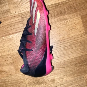 Adidas x speedportal - Säljer ett par snygga fotbollsskor från Adidas i rosa och svart med silvriga detaljer. Skorna har snörning och en smal passform, perfekt för dig som vill sticka ut på planen. Yttermaterialet är syntet och sulan är platt med dobbar.