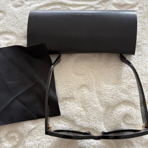 Svarta solglasögon från Saint Laurent - Stilrena svarta solglasögon från Saint Laurent med rektangulära bågar och guldigt YSL-logo på sidorna. Bågarna är i blank plast och har en klassisk, tidlös design. Kommer med originalfodral och putsduk.