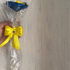 Blå och gul ros i satin med rosett - En handgjord ros i blått och gult satinband, inslagen i cellofan och dekorerad med en gul rosett med blå sten. Perfekt som accessoar till student eller midsommar.