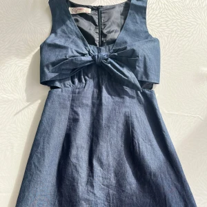 Mörkblå ärmlös klänning med knytdetalj från F Fashion Paris - Supersnygg mörkblå jeansklänning från F Fashion Paris med knytdetalj framtill och markerad midja. Klänningen är ärmlös och har en klassisk rund halsringning samt dragkedja i ryggen. 