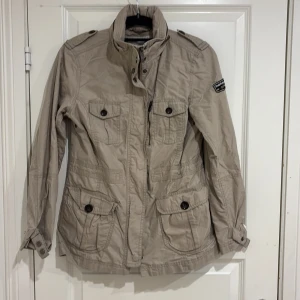 Beige jacka från Peak Performance - Snygg beige jacka från Peak Performance med flera stora fickor framtill och knappar. Jackan har hög krage, axelklaffar och ett broderat märke på ena ärmen. Perfekt för vår och höst, med lång ärm och klassisk design.