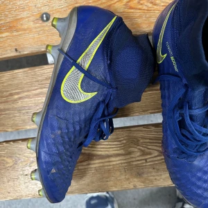 Blå Nike fotbollsskor med högt skaft - Säljer ett par blå Nike fotbollsskor med högt skaft och snörning. Skorna har en stor gul Nike-logga på sidan och är tillverkade i syntetmaterial med struktur för bättre bollkontroll. Sulan har fasta dobbar för spel på gräs. Nike magista obra 2 (väldigt sällsynta)