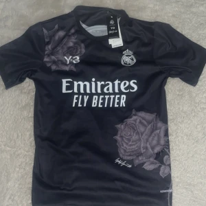Real Madrid svart fotbollströja Y-3 x Adidas - Svart Real Madrid fotbollströja i samarbete med Y-3 och Adidas. Tröjan har korta ärmar, stora ros-motiv på bröstet och nedre delen samt klubbmärke och sponsortryck på framsidan. Materialet är lätt och andas, perfekt för match eller träning. Storlek S men passar perfekt för M också 