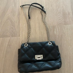 Svart axelväska med kedja - Säljer en svart  axelväska med silverfärgad kedjerem och snyggt spänne. Insidan har dragkedja för extra säkerhet. Stilren och enkel att matcha.