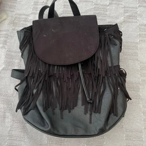 Svart mocka ryggsäck med fransar - Säljer en svart ryggsäck med långa fransar framtill. Väskan har dragskostängning och ett lock. Tillverkad i syntetmaterial med justerbara axelremmar. Perfekt för dig som vill ha en cool och trendig accessoar.