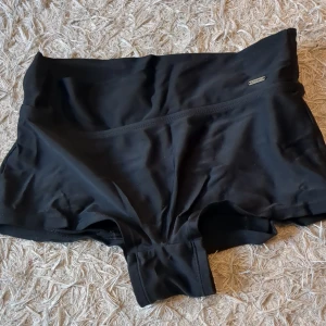 Svarta badshorts - Svarta, korta badshorts från Firefly med bred resår i midjan. Tillverkade i ett stretchigt syntetmaterial som passar perfekt för simning eller strandhäng. Tyvärr har materialet blivit lite konstigt av sanden på stranden men det är inte som syns när man har på sig dem och man kan vika ner dem så dom blir lågmidjade.
