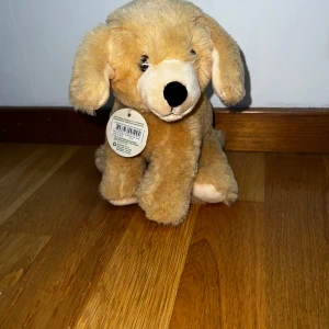 Gosedjur hund mjukisdjur - Säljer ett gulligt mjukisdjur i form av en hund. Hunden är ljusbrun med mörk nos och svarta broderade ögon. Pälsen är mjuk och fluffig, och den har långa öron och en liten svans. Perfekt som kramkompis eller dekoration. Helt ny med lapp kvar. Inte använd alls