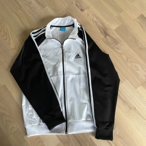 Svartvit träningsjacka från Adidas - Säljer en klassisk svartvit träningsjacka från Adidas med dragkedja och ståkrage. Jackan har svarta ärmar, vita paneler framtill och bak samt de ikoniska Adidas-ränderna längs axlarna. Perfekt för sport eller chill.