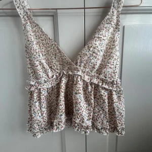 Blommig volangtopp med v-ringning - Supersöt blommig topp med volangdetaljer och v-ringning. Toppen är ärmlös och har smock i ryggen för en bekväm passform. Perfekt för varma dagar och somriga outfits.