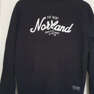 Svart hoodie från Norrland - Svart hoodie med vit text 'The Great Norrland' på bröstet. Klassisk känguruficka framtill och huva med dragsko. Perfekt för dig som gillar enkel och stilren streetwear.