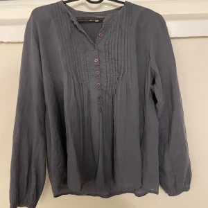 Grå blus  - Säljer en grå blus från Rabalder med dekorativa veck framtill och rosa knappar. Blusen har lång ärm och ett broderat motiv med krona på ena sidan. Perfekt för dig som gillar klassiska detaljer.