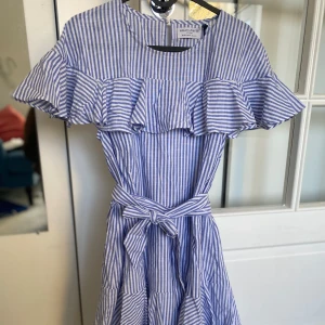 Blåvit randig klänning med volang  - Supersöt blåvit randig klänning från Holly & White med volangdetalj över axlarna och knytskärp i midjan. Perfekt för vår och sommar! Rund halsringning och kort ärm.