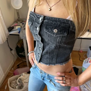 Sommar tröja - Säljer en svart croppad tubtopp i jeanstyg med knappdetaljer och bröstfickor framtill. Toppen har en smockad baksida för extra komfort och sitter snyggt tight. Perfekt för dig som vill ha en unik och trendig look. Faktiskt jätte skön🥰 Xs/ S