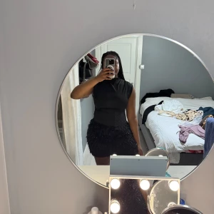 Svart kortklänning med en kjol  - Säljer en svart kortklänning med tight passform och ärmlös design. Klänningen har en slät ovandel och kjolen är dekorerad med svarta fjädrar som ger en unik och trendig look. Perfekt för dig som vill sticka ut på festen!