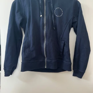 Mörkblå hoodie med dragkedja från Marine - Mörkblå hoodie från Marine med dragkedja framtill och huva. Tröjan har två fickor på sidorna och en liten vit logga på bröstet. Perfekt för en avslappnad stil.