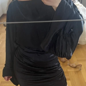 Svart kort klänning med vida ärmar - Säljer en svart kort klänning med glansig finish och markerad midja. Klänningen har vida, långa ärmar och en lätt rynkad kjol som ger en snygg siluett. Perfekt för dig som vill sticka ut med en elegant och trendig look. Klänningen är använd så lite sliten, men inget som påverkar funktionen. 