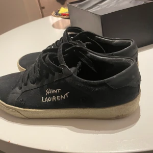 Svarta Saint Laurent sneakers - Svarta sneakers från Saint Laurent med vit logga broderad på sidan. Skorna har svart snörning, rund tå och vit platt sula. Ovandelen är i canvas och detaljer i skinn. Perfekt för dig som gillar en stilren men edgy look. Kan gå ner priset vid snabb affär,kontakta vid frågor 100% äkta bok påse kuvert finns, inget kvitto, köpte d från plick