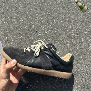 Maison Margiela - Säljer ett par svarta Maison Margiela sneakers med beige sula och vita snören. Skorna har en mix av mocka och skinn, klassisk design och rund tå. Perfekta för dig som gillar stilrena och tidlösa sneakers.