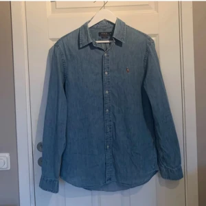 Ljusblå jeansskjorta från Polo Ralph Lauren - Säljer en klassisk ljusblå jeansskjorta från Polo Ralph Lauren med broderad logga på bröstet. Skjortan har lång ärm, knappar framtill och en avslappnad slim fit-passform. Perfekt till många olika outfits!