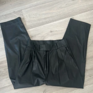 Svarta kostymbyxor i skinnimitation - Snygga svarta kostymbyxor i skinnimitation med hög midja och pressveck. Byxorna har raka ben och hällor för bälte. Perfekta för en trendig och stilren look.