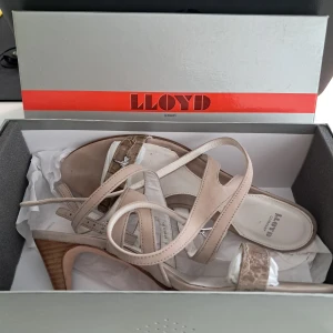 Beige sandaler med klack från Lloyd - Säljer ett par eleganta beige sandaler från Lloyd med smala remmar och öppen tå. Skorna har en medelhög klack i trälook och en dekorativ detalj framtill. Perfekta för sommarens tillfällen!