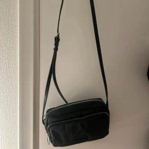 Svart axelväska från Pull&Bear - Säljer en stilren svart axelväska från Pull&Bear med justerbart axelband och dragkedja. Aldrig använd 