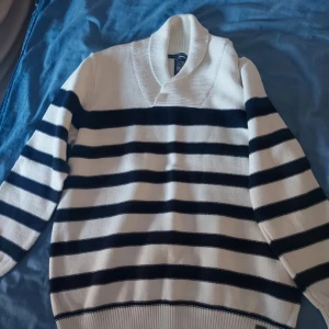 Vit och marinblå randig stickad tröja - Säljer en vit stickad tröja med marinblå horisontella ränder och bred krage. Tröjan har långa ärmar och ribbade muddar vid ärmslut och nederkant. Perfekt för kyliga dagar och en klassisk stil.