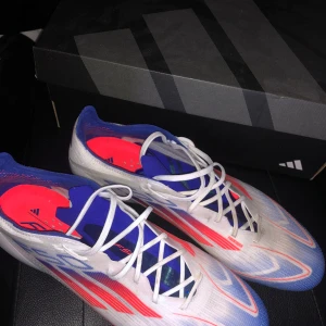 Adidas X Speedflow fotbollsskor - Säljer ett par Adidas F50 Elite använda 2-3 ggr  fotbollsskor i vitt med blå och röda detaljer. Skorna har snörning och är designade för fart på planen, med en lätt ovandel och fasta dobbar för bra grepp. Perfekta för dig som vill sticka ut på fotbollsplanen.
