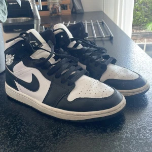 Nike Air Jordans svartvita sneakers - Säljer ett par klassiska svartvita Nike Air Jordans med högt skaft och svart snörning. Skorna har vit bas med svarta detaljer och den ikoniska Air Jordan-loggan på sidan. Perfekta för dig som gillar streetwear och vill ha en tidlös look.