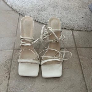 Vita klackade sandaler med snörning - Säljer ett par vita sandaler med hög klack och fyrkantig tå. Skorna har tunna remmar och lång snörning som kan lindas runt vristen. Perfekta för dig som gillar stilrena och trendiga skor.
