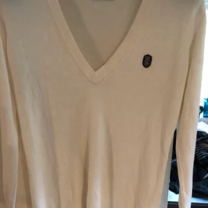 Vit v-ringad tröja från Gina Tricot - Säljer en vit v-ringad tröja från Gina Tricot i storlek XL. Tröjan har lång ärm och en liten svart broderad logga på bröstet. Perfekt basplagg till garderoben!