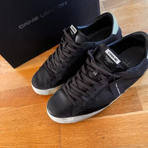 Svarta sneakers från Crime London - Säljer ett par svarta sneakers från Crime London med vit sula och svarta skosnören. Skorna har en klassisk låg modell och är tillverkade i skinn med diskreta detaljer. Perfekta för en stilren vardagslook.