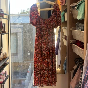 Blommig långklänning med puffärm - Säljer en långklänning med blommigt mönster i rosa, rött och orange på mörk botten. Klänningen har puffärmar och smockad överdel som ger en snygg passform. Perfekt för sommarens alla tillfällen! 